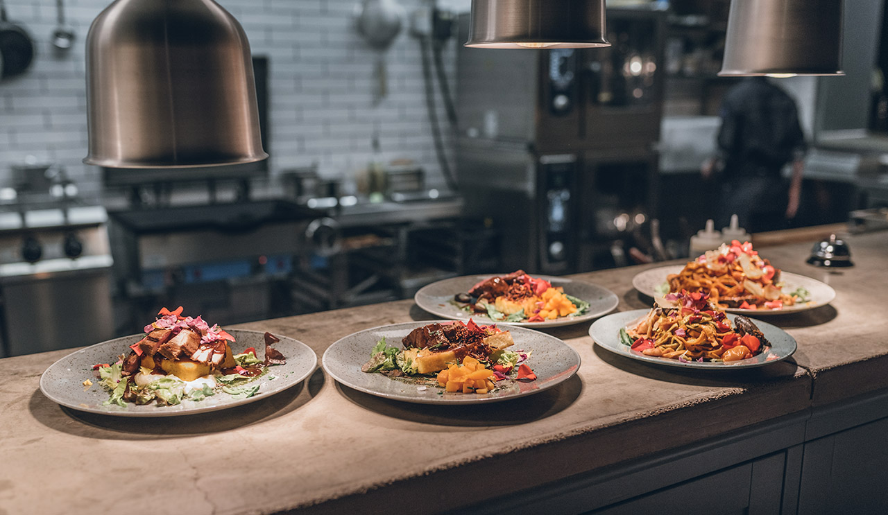 Fem tallrikar med gourmetmat är arrangerade på en bänk under varma pendellampor i ett professionellt kök, med en kock suddig i bakgrunden och redskap synliga på hyllorna.