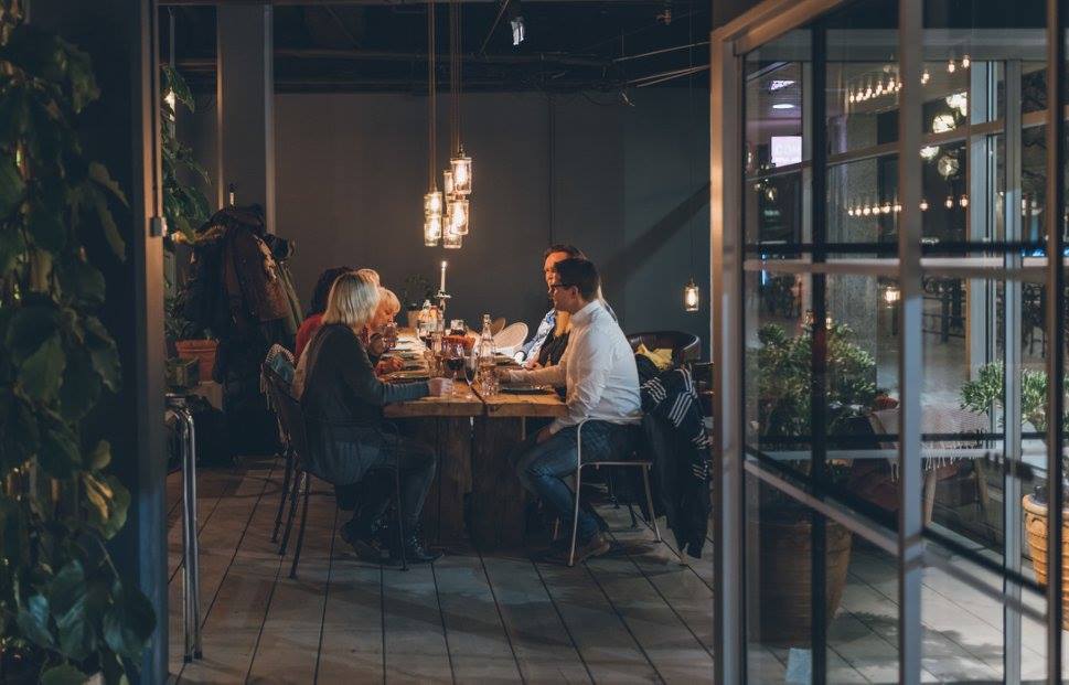 En grupp människor sitter runt ett träbord och avnjuter en måltid tillsammans i en mysig, varmt upplyst restaurang med hängande lampor och krukväxter som syns genom stora glasfönster.